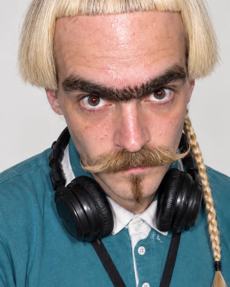 Suno with a blond bowl cut, heavy unibrow, mustache, braid, headphones and teal polo.